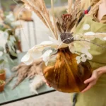 Floral designer holding a large amber vase with an elegant dried arrangement of pampas grass, eucalyptus, and seed pods, showcasing a seasonal botanical display for estate garden enhancements.