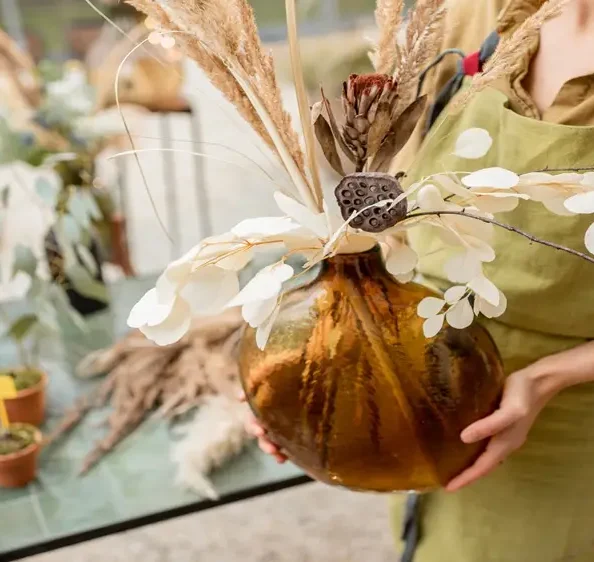 Floral designer holding a large amber vase with an elegant dried arrangement of pampas grass, eucalyptus, and seed pods, showcasing a seasonal botanical display for estate garden enhancements.