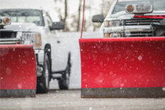 Two pickup trucks with red snow plows parked and ready during active snowfall.